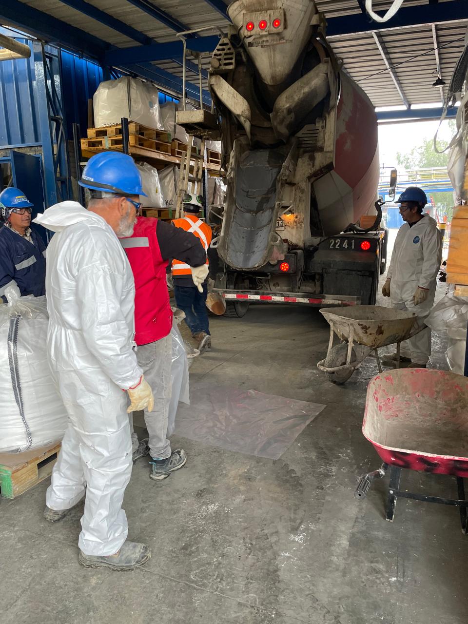 Operarios con trajes de seguridad y cascos supervisan la descarga de un camión mixer en un galpón industrial, preparando la aplicación de morteros de reparación.