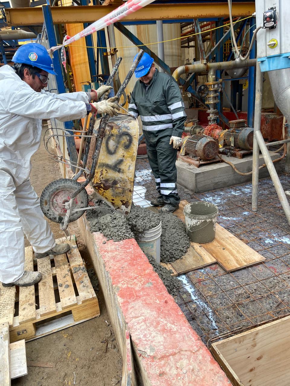 Operarios industriales vertiendo mortero de reparación desde una carretilla sobre una malla de refuerzo en una zona de contención de planta industrial.