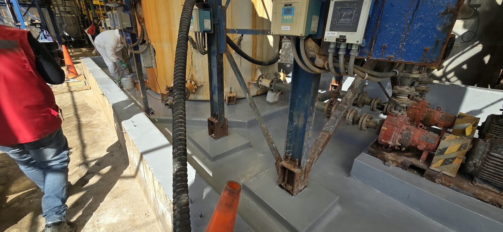 Pretil de contención de hormigón en planta industrial, recubierto con una capa polimérica gris lisa para protección química, con bombas y estructuras de soporte.