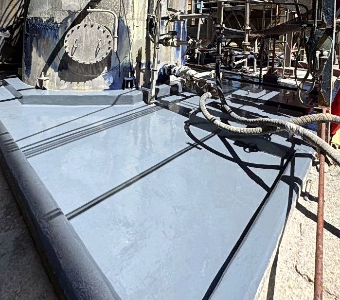 Base de hormigón y pretil de contención secundario con recubrimiento polimérico gris aplicado junto a estanque industrial y tuberías.