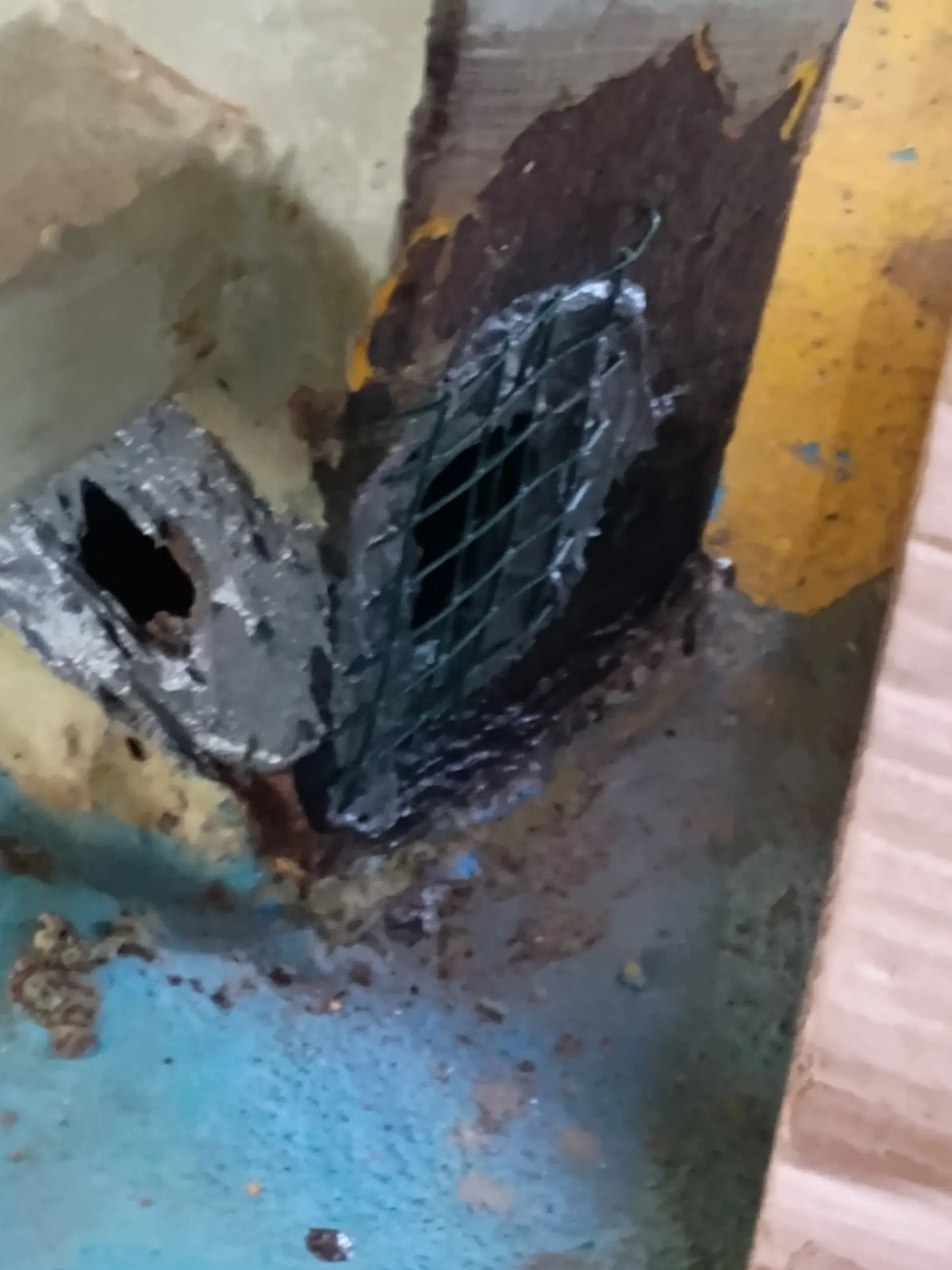 Estructura metálica severamente corroída con perforación grave, parcheada con malla de refuerzo y compuesto de reconstrucción polimérico gris durante su reparación.