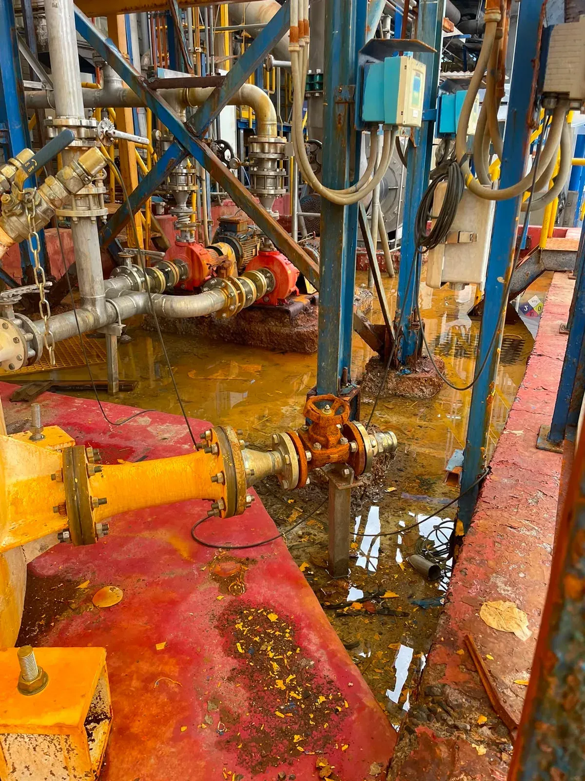 Piso industrial y estructuras metálicas con severa corrosión y acumulación de fluidos corrosivos en una planta de procesos, mostrando degradación del recubrimiento protector.