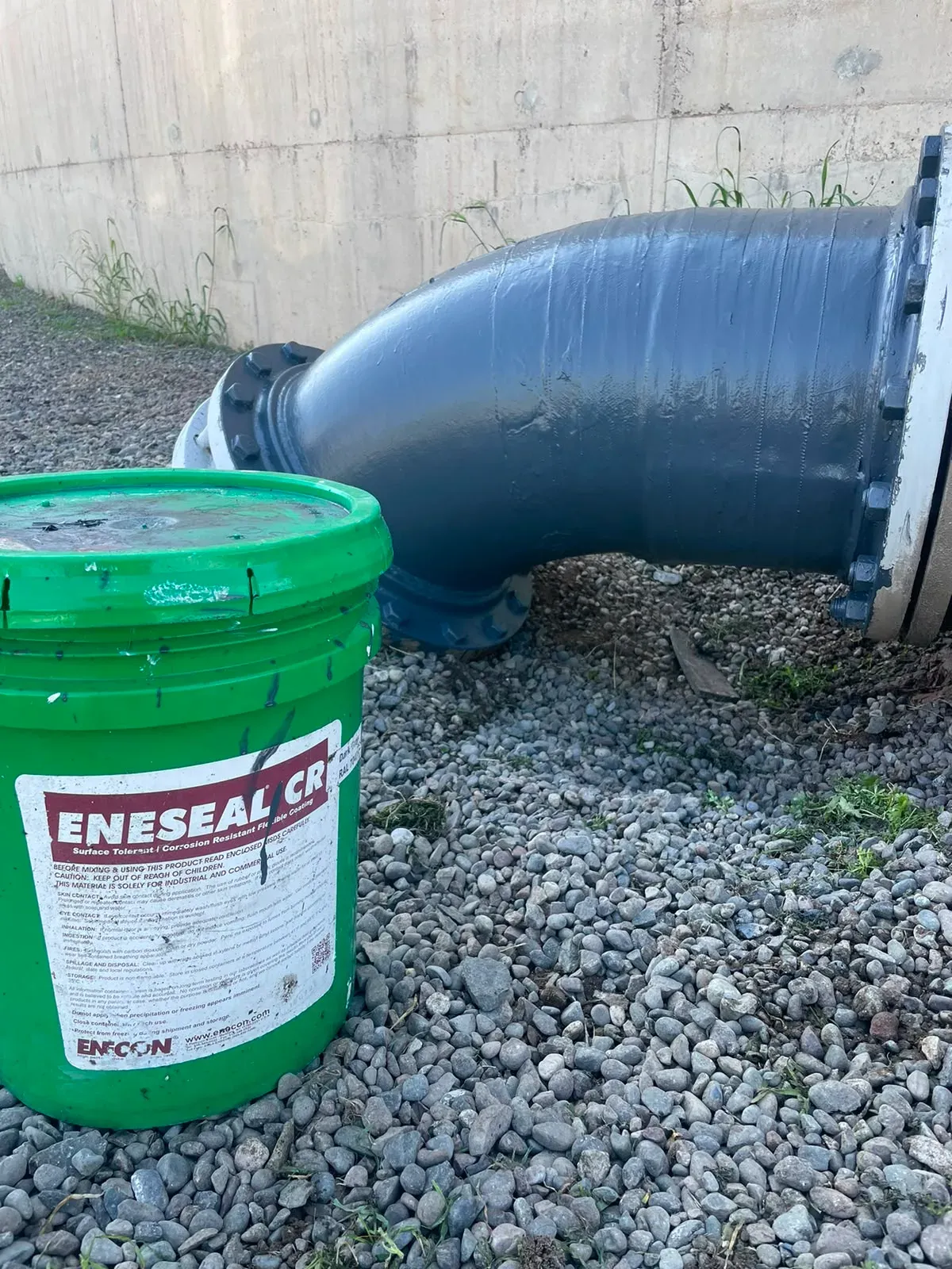 Balde de ENESEAL CR en primer plano junto a un codo de tubería metálica protegido con recubrimiento polimérico gris oscuro contra la corrosión.